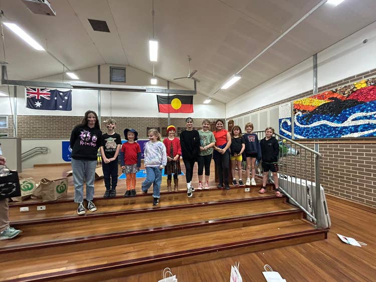 A teacher and group of students of all ages on the stage in the school hall participating in a group activity.